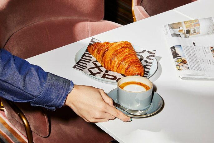 Hand reaching for a cup of coffee on a table, with a croissant on a plate and newspaper nearby.