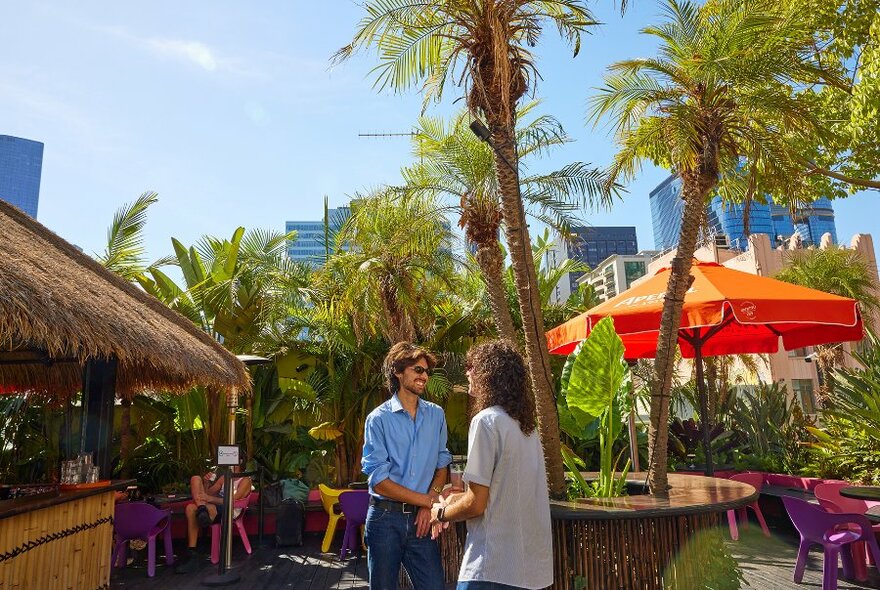 Two people chatting on a rooftop bar with palm trees, orange umbrellas and a thatched roof bar. 