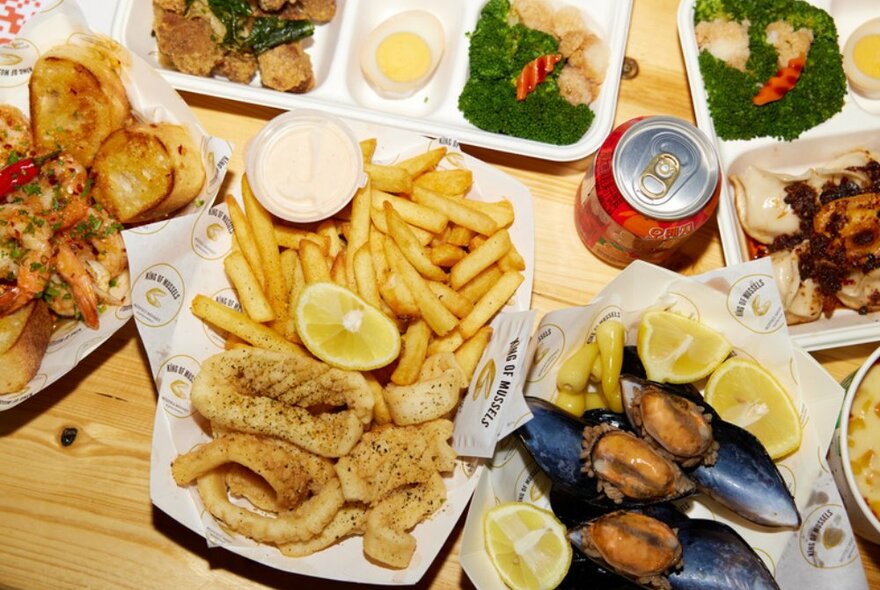 Birds-eye view shot of food on wooden table. Next to a can of coke is fish and chips, bento boxes, and dumplings.