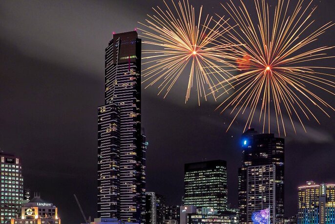 The Eureka Tower and other city skyscrapers, with two large orange starburst fireworks in the night sky behind.