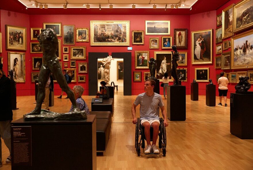 A gallery space with red walls covered in paintings and statues throughout. A person is moving through the room using a wheelchair.