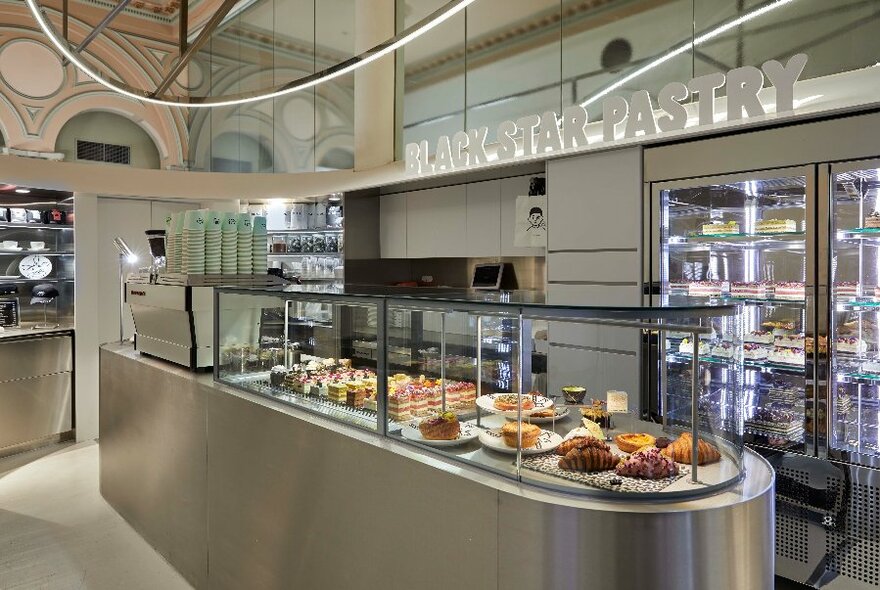 Slick steel and glass interior of a cafe with plates of cakes and pastries on display behind a glass counter, a coffee machine also visible.