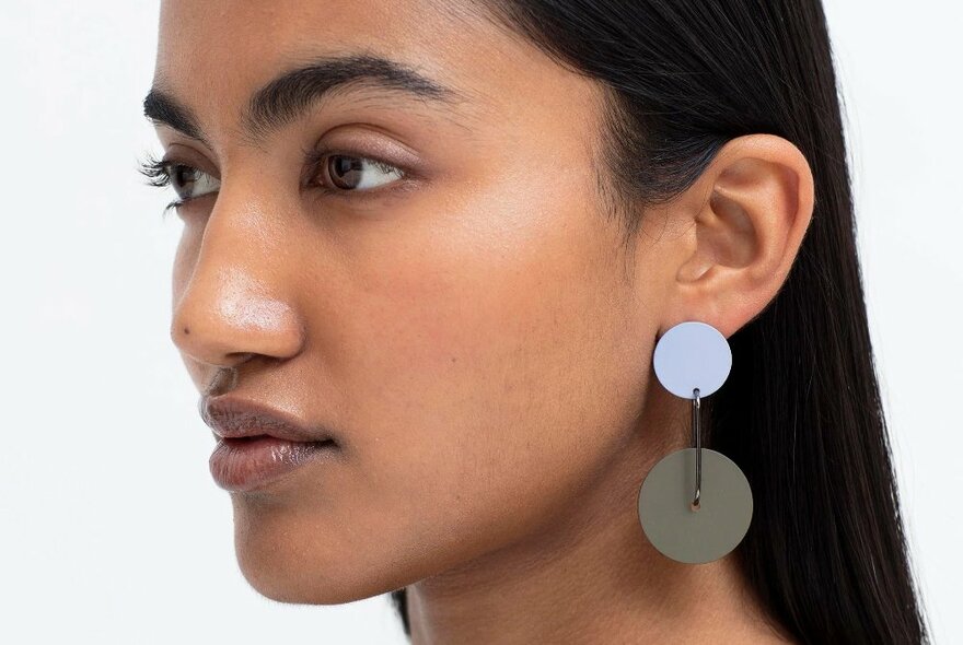 A close up shot of a model wearing earrings with a light blue circular stud and a larger grey circle hanging below.