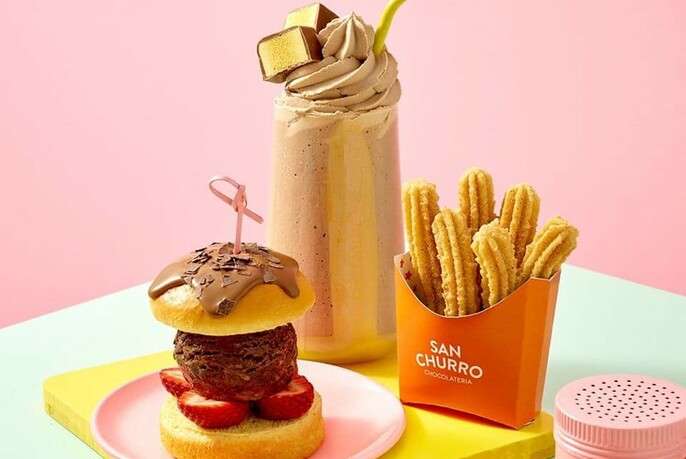 Pink background with green table in foreground with packet of churros, chocolate ice cream burger and chocolate milkshake with ice cream and chocolate honeycomb garnish.