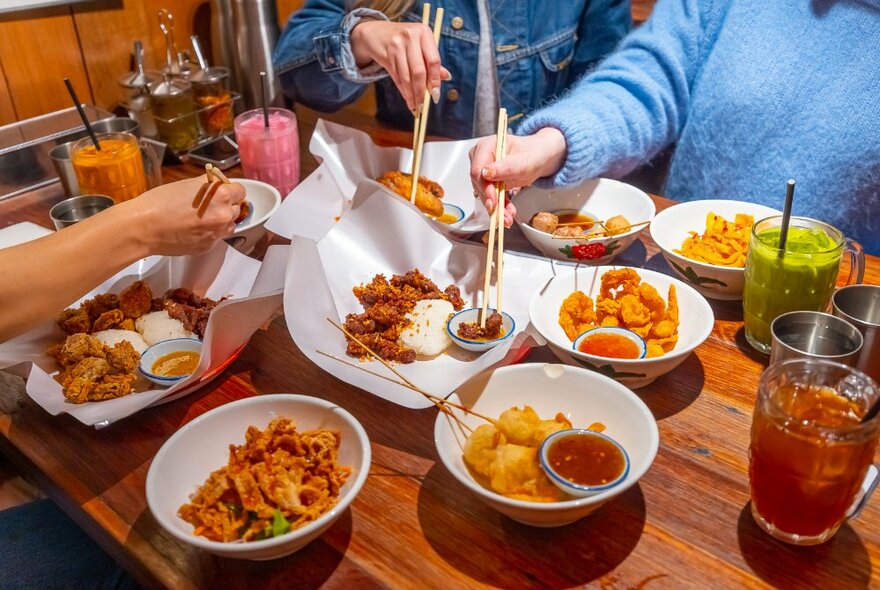 Three sets of hands using chopsticks to pick up food from plates on a table, colourful drinks also on the table.