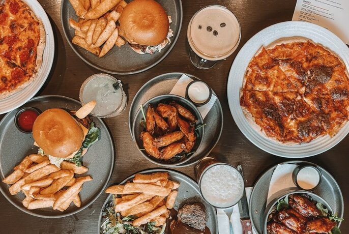 Overhead view of plates of food on a table including pizza, burgers and fries, chicken wings and glasses of beer.