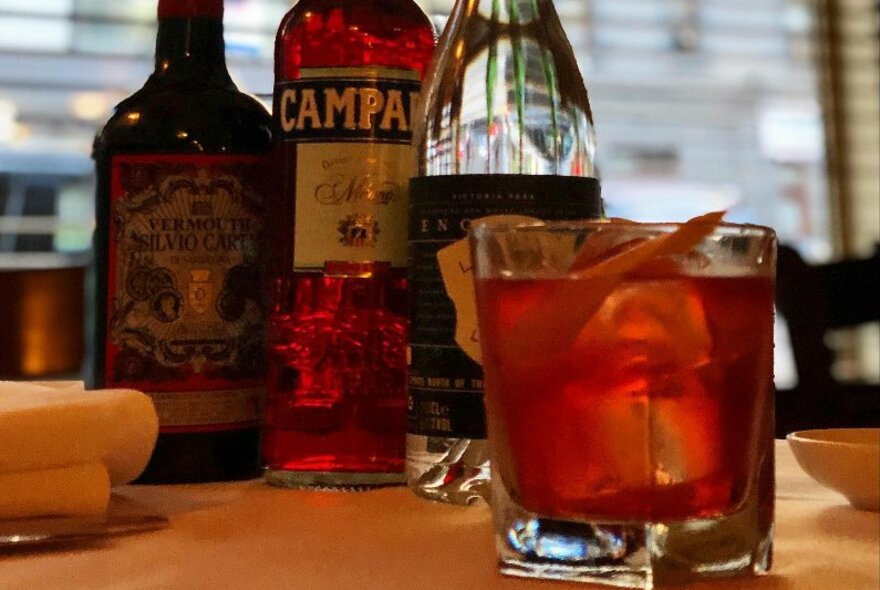 A negroni cocktail and the three bottles that make up the ingredients in the background.