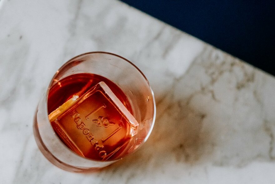 Looking down on a campari-based cocktail with a single square ice cube on a marble tabletop. 