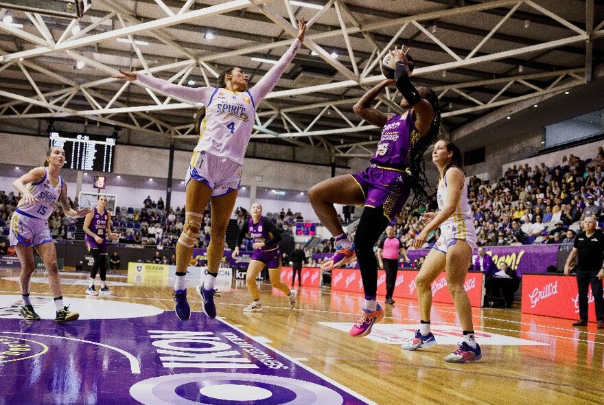 Women playing basketball on a court, two players from opposing teams jumping high in the air, with a large stadium crowd in the background.