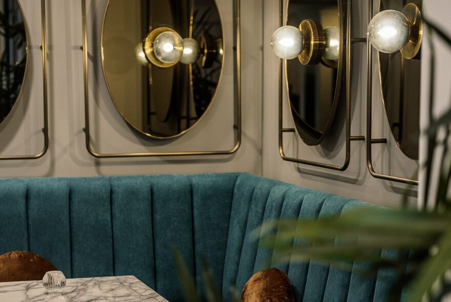 Interior of a bar with blue-green velvet booth seating, art deco style round mirrors on the wall and soft pearly lamps.