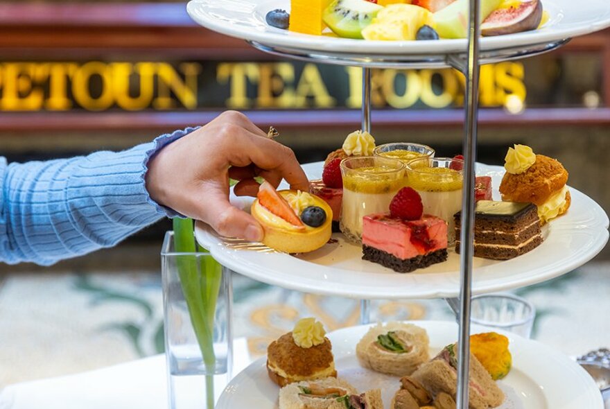 Someone reaching for a lemon tart on a high tea tiered stand full of bite sized treats.