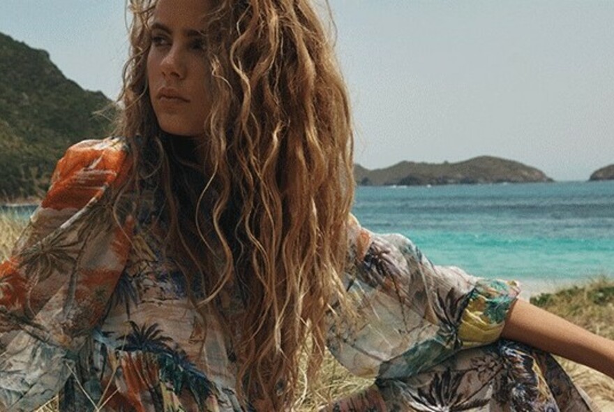 Model with long, wavy hair on a beach wearing a Zimmermann ocean-patterned dress.