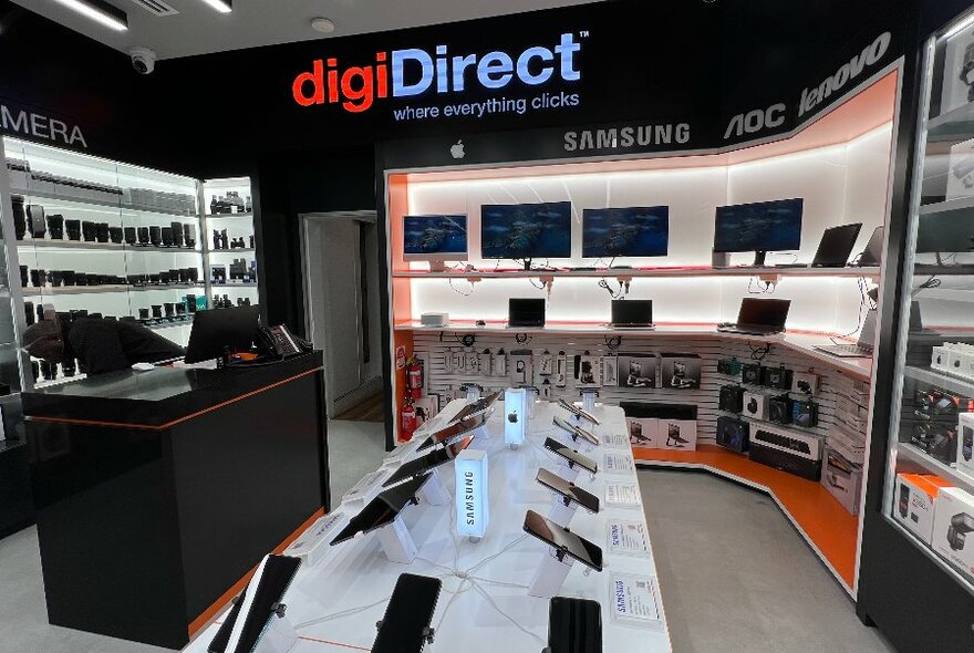 Digital store interior with wall signage and backlit white shelves displaying screens and other technological products.