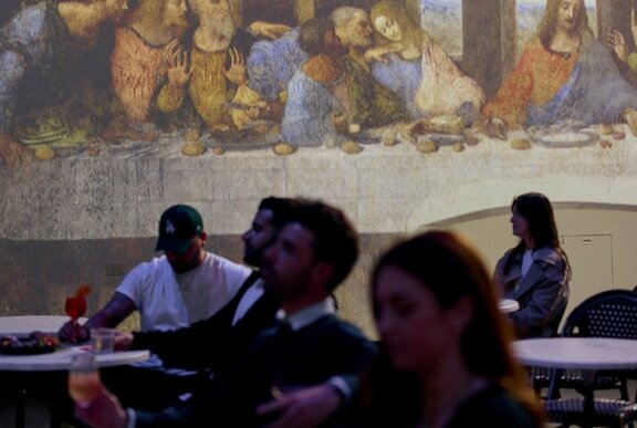 People seated dining and drinking at tables with Leonardo's painting of the Last Supper projected on a wall behind them.