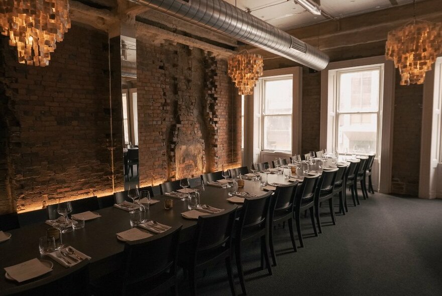 A long formal dining table set for dinner in a dimly lit room with dark walls and moody fabric chandeliers.