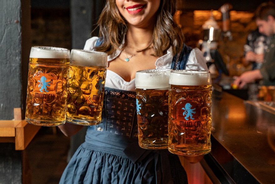 A person wearing a traditional Oktoberfest dress holding four large steins of beer in her hands.