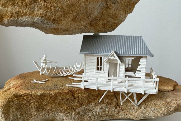 Sculpture of a small house made from small bits of matchstick like wood, nestled between two large rocks.