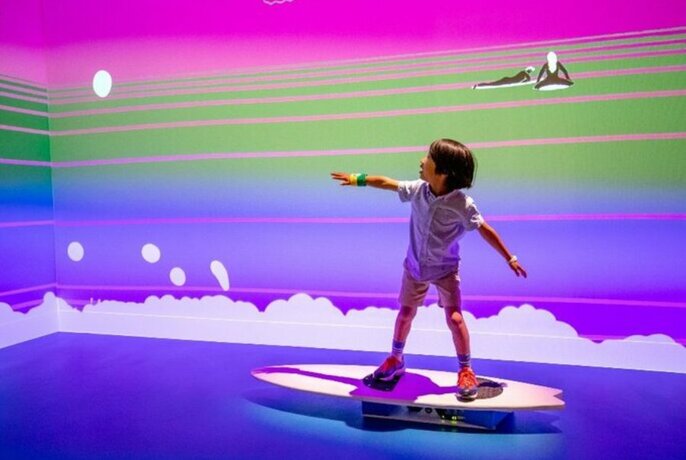 Young child balanced on surfboard in a room painted to look like the ocean.