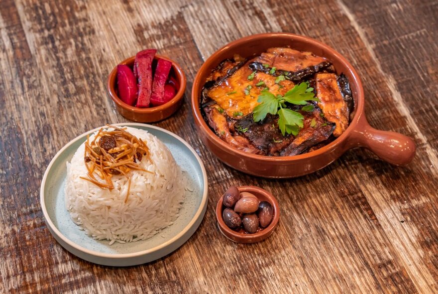 Food in small dishes, including rice, pickled beets, olives and a small tagine of eggplant, resting on a wooden table