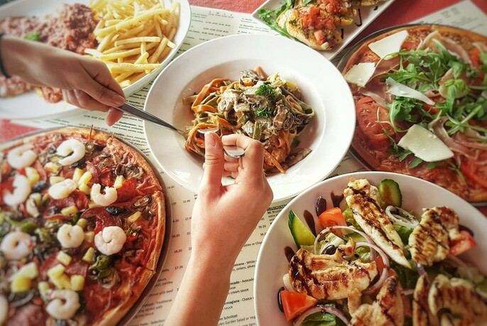 Hands selecting food from plates of food on a table, including salads, pizza and chips.