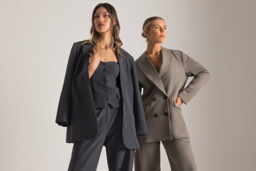 Two female models wearing fashionable business style suit jackets and loose trousers, posing against a light coloured wall.