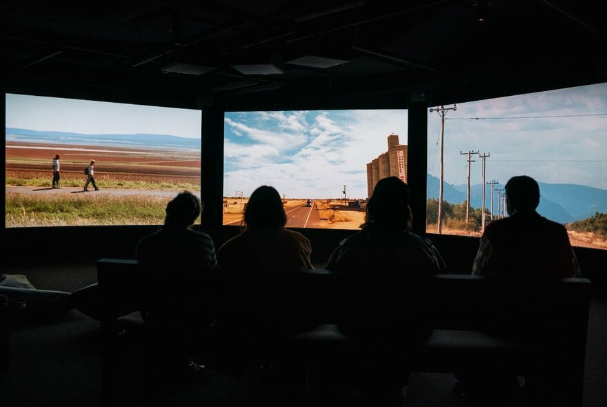 Three large screens side-by-side showing outback scenes and the silhouettes of people watching in front.