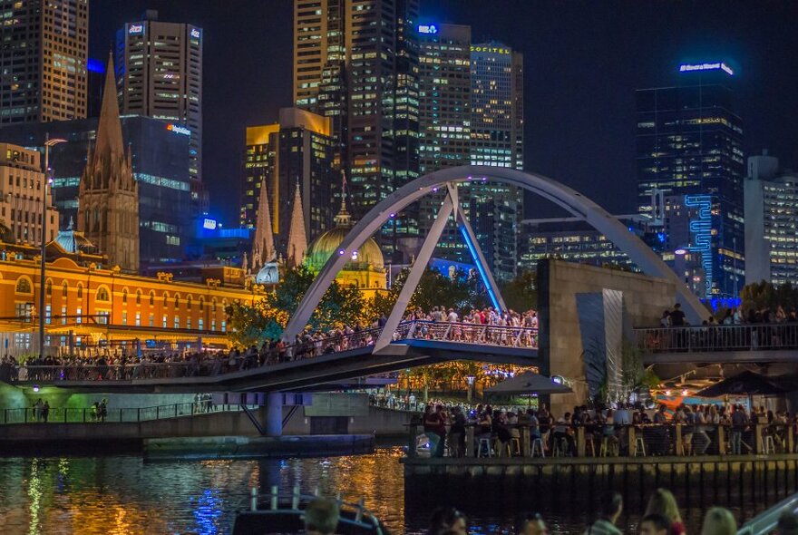 Crowds of people on a bridge and along the banks of the Yarra River with illuminated city building in the background.
