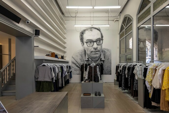 Fashion garments on racks in a shop, with a large black and white print of a man's head in background.