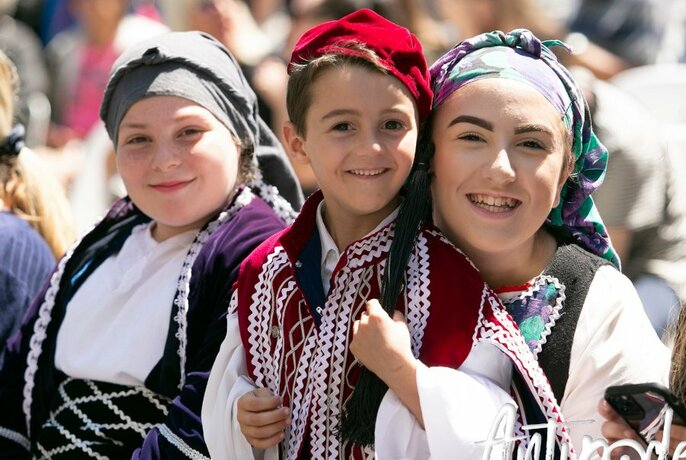 Three young people in Greek national dress, smiling. 