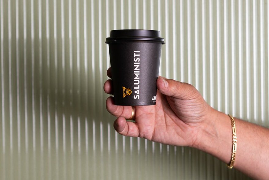 A hand holding a Saluministi takeaway coffee in front of a cream-coloured fluted wall.