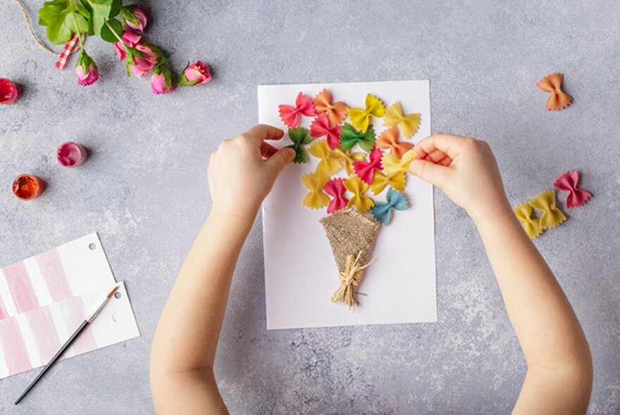 Overhead view of the arms of a child making a craft card using dried flowers, colourful butterfly-shaped pasta and paint.