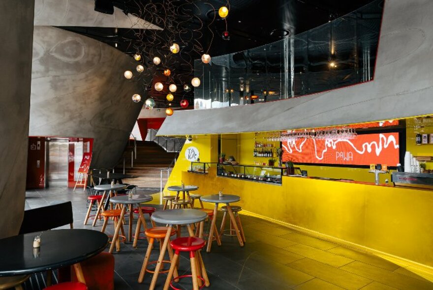 A bright yellow interior of a cafe with small tables and red stools. 