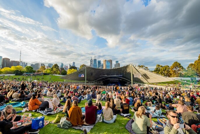 Groups of people sitting on rugs on the lawn in front of the Sidney Myer Music Bowl structure, with the Melbourne city skyline visible in the far background.