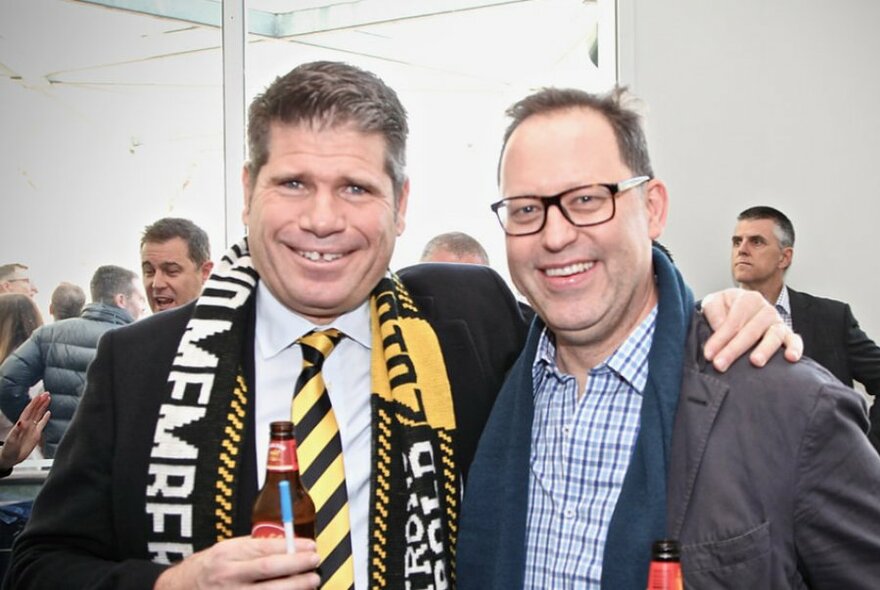 Two smiling men wearing suits and with their arms around each other's shoulders, one of them wearing a team scarf and holding a small beer bottle. 