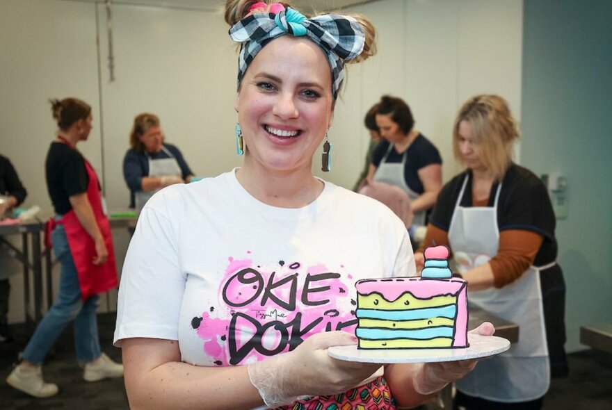A woman smiling as she holds a slice of illustrated cake on a plate, a large checked bow in her hair. 