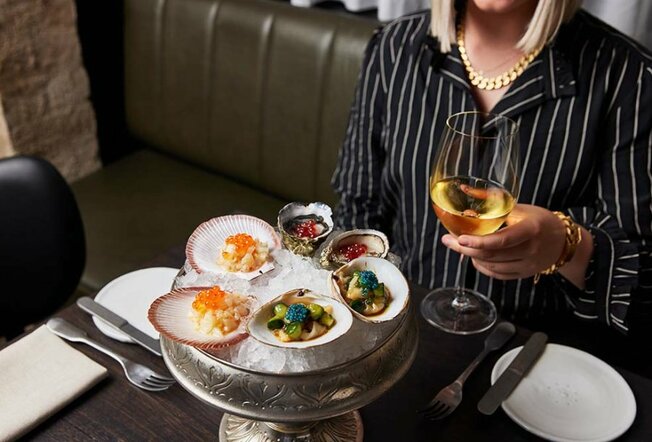 A woman holding a glass of white wine with a tray of oysters topped with colourful garnishes.