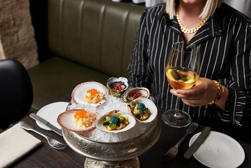 A woman holding a glass of white wine with a tray of oysters topped with colourful garnishes.