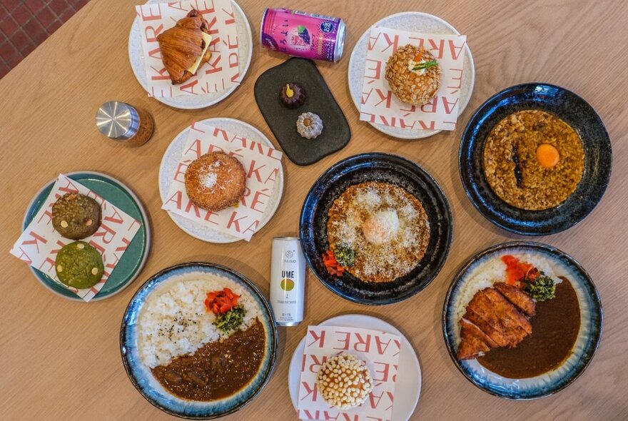 Overhead view of a table filled with various plates of Japanese food including curry, rice, curry pan and mochi sweets, plus cans of drink.