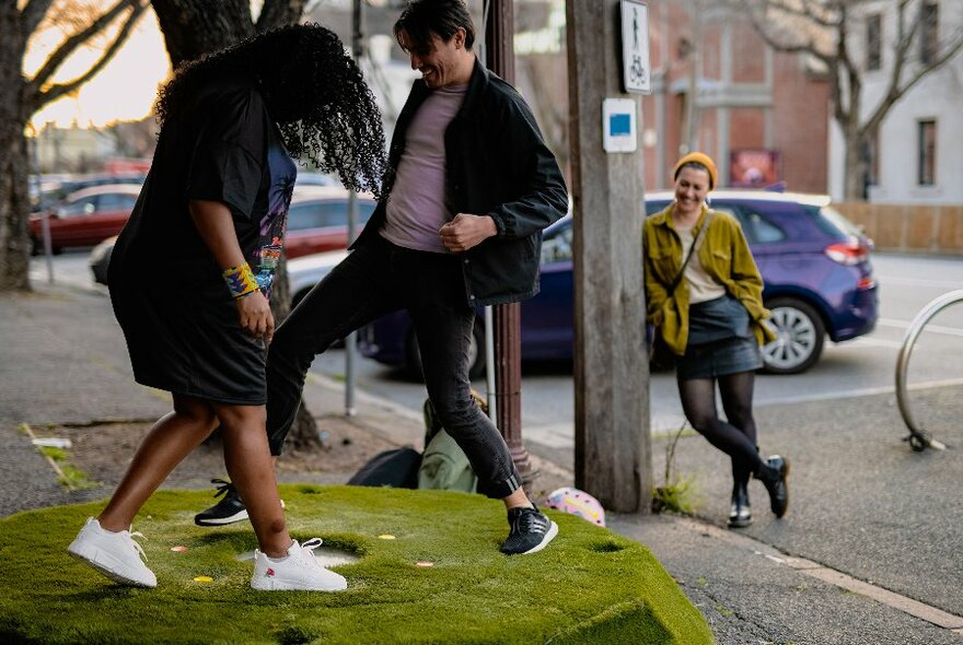 People playing a game on a nature strip with another person leaning on a pole watching them. 