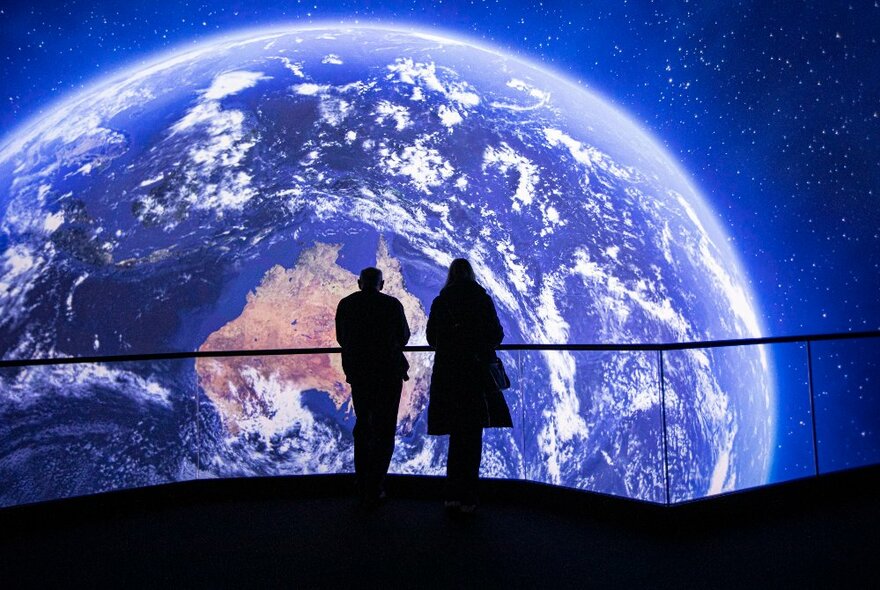Shadows of two people silhouetted in front of a huge digital projection of the earth in space, with the continent of Australia visible.