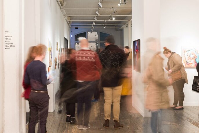 Blurred image of people moving through a gallery space with artworks on the walls.