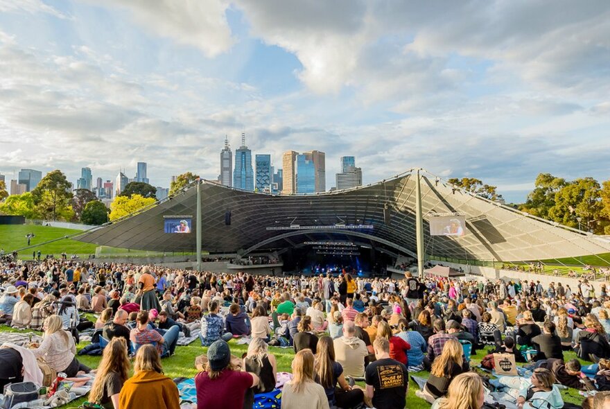 Large crowd of people seated on the grass reserve in front of the Sidney Myer Music Bowl arena, with the Melbourne city skyline of buildings in the far background.