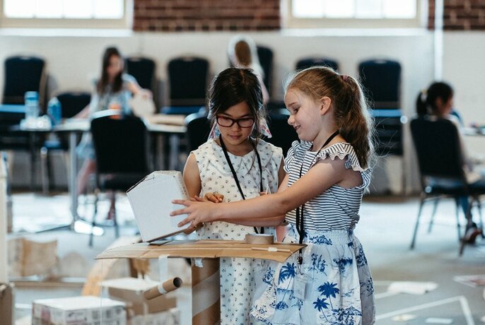 Two girls aged about 8 or 9 working together to create a cardboard structure in a large open space, with chairs, tables and other young children in the background.