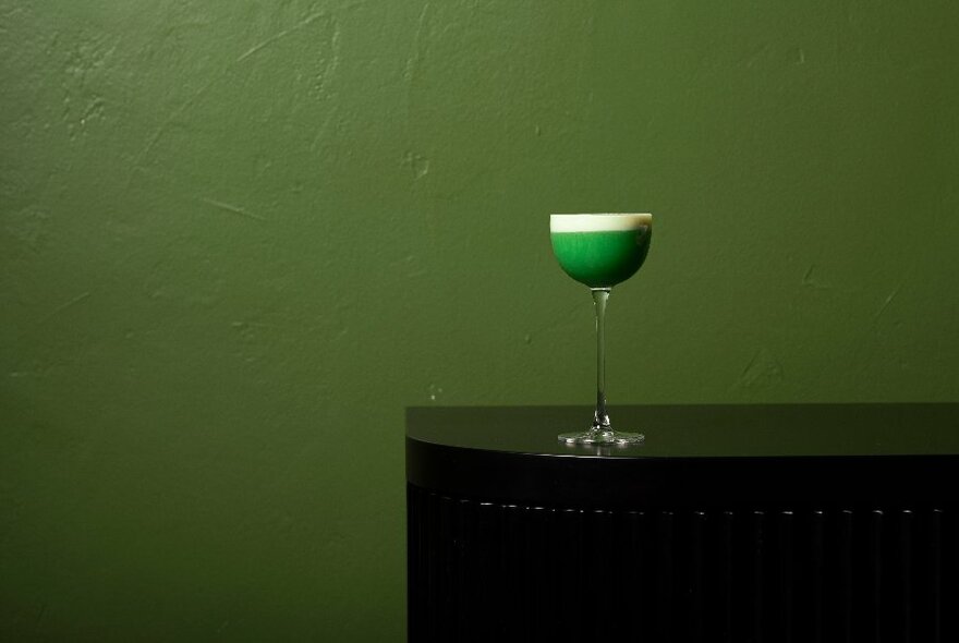 A bright green foamy cocktail in front of a green wall. 
