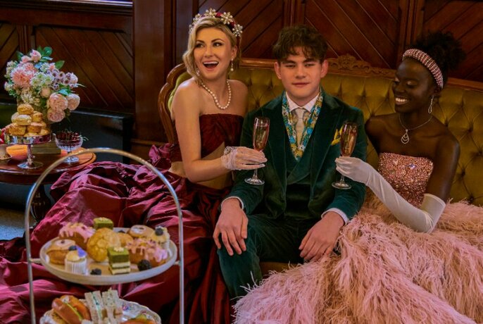 Three people reclining on a velvet couch, dressed in Regency inspired gowns and a velvet suit, with plates pf food in front of them.