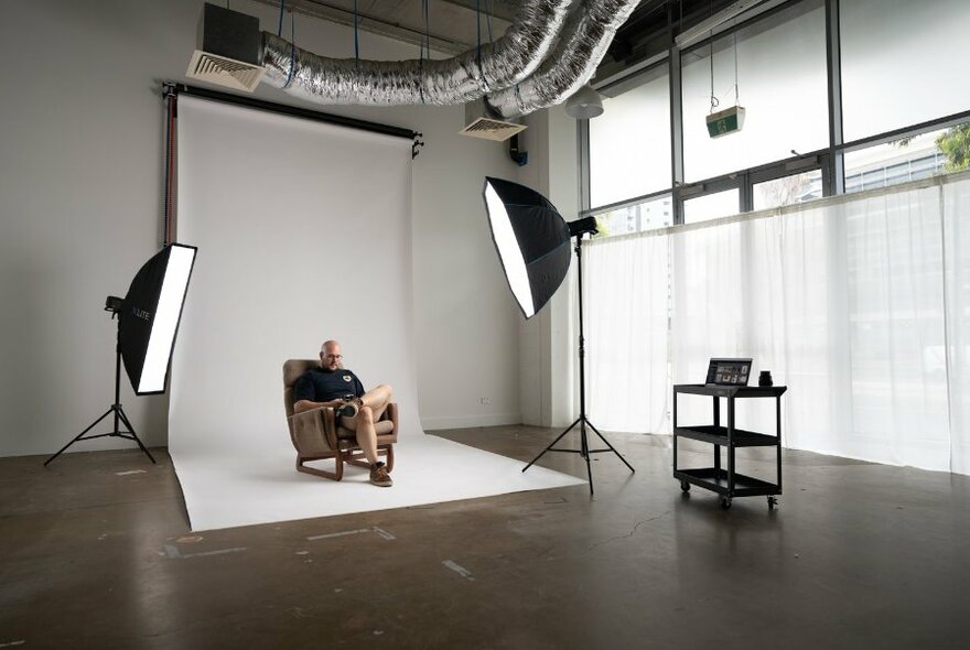 A photo shoot in a large studio space, with a person sitting in an armchair against a white screen backdrop, two large camera lights angled towards them.