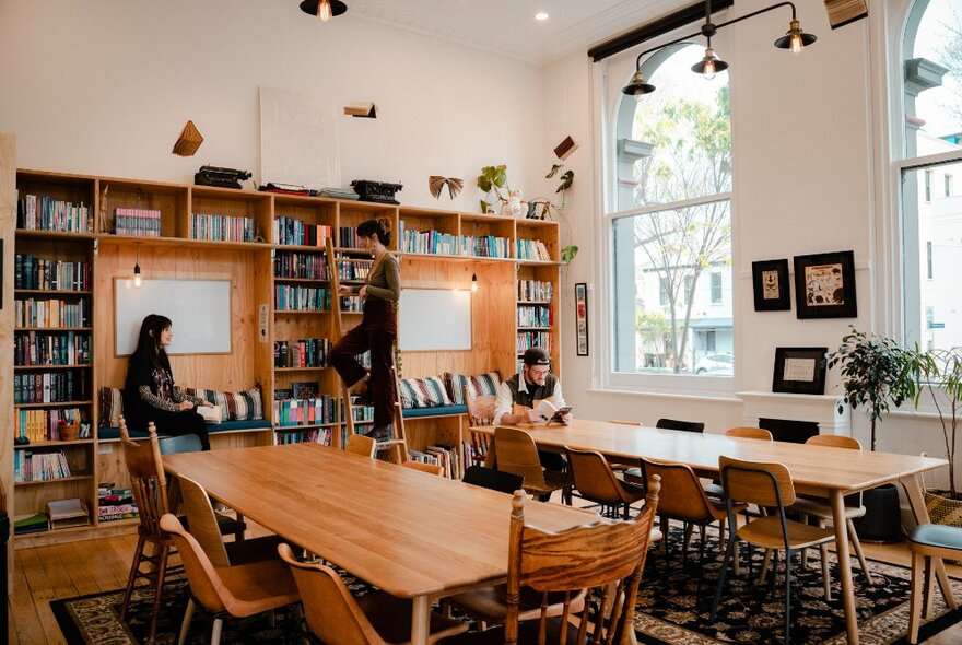 A workshop room with bookshelves and large wooden tables with chairs in a light naturally lit room with large windows.