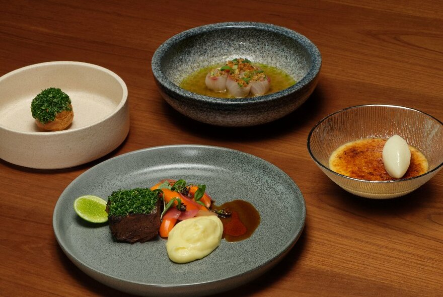 Four dishes of carefully arranged restaurant-styled food on ceramic plates and bowls on a  wooden table.