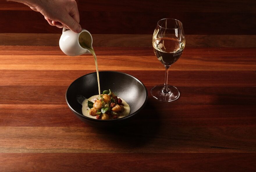 A hand pouring a jug of white sauce into a dark dish of food, with a wine glass on a wooden table.