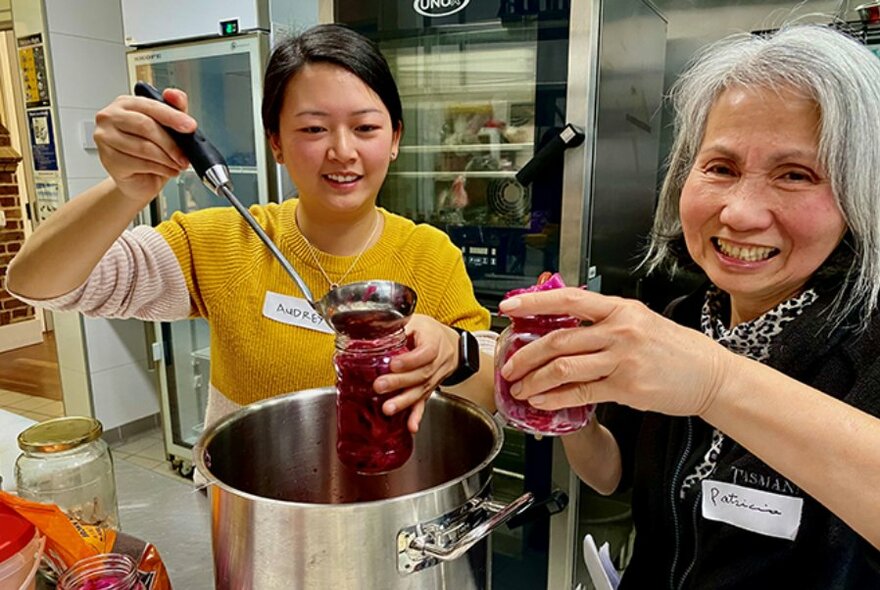 Two smiling women in a commercial kitchen bottling up red pickles, one using a ladle. 